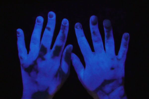 Mit UV-Licht beleuchtete Hände, auf denen Benetzungslücken bei der Händehygiene sichtbar werden.